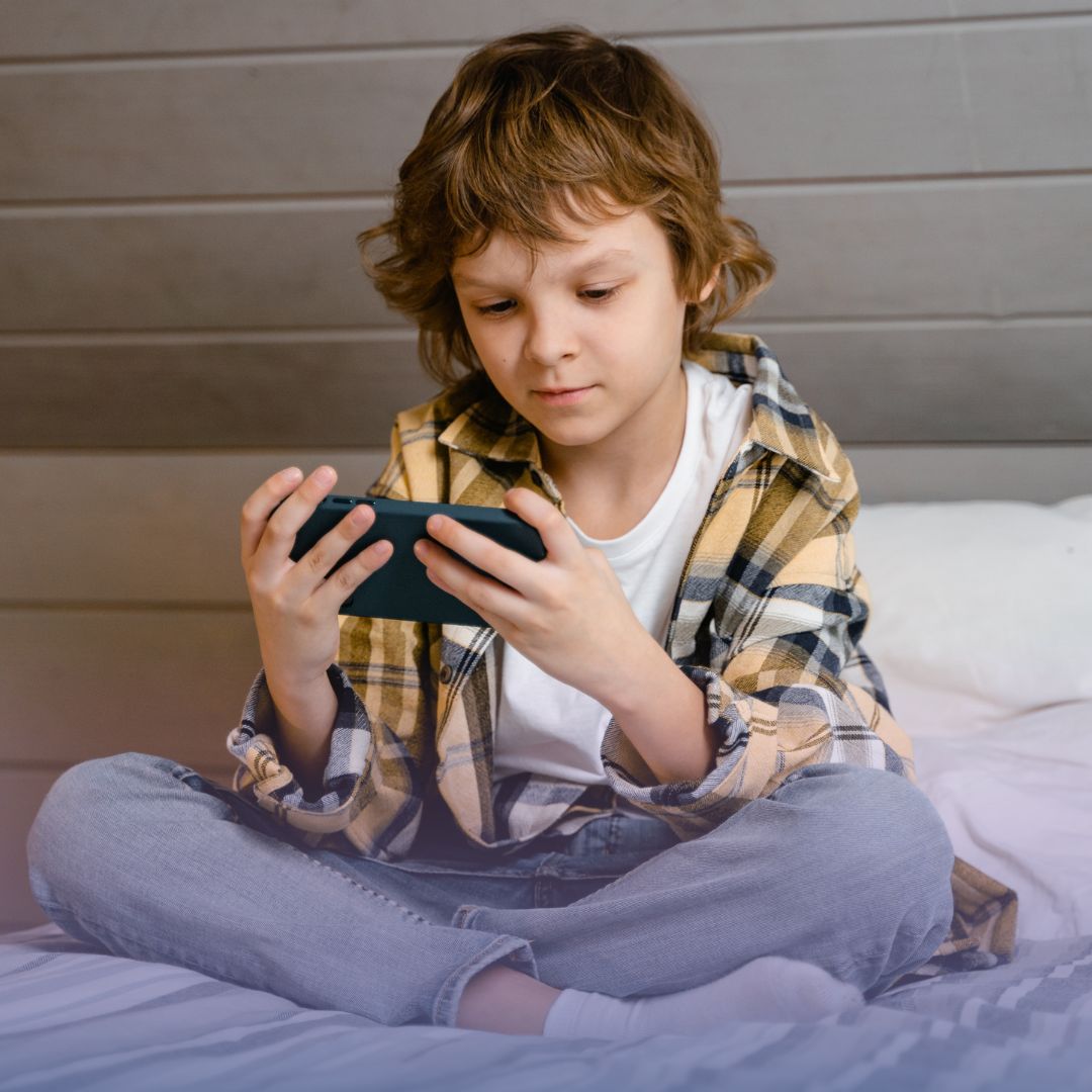 Criança sentada na cama olhando para o celular, ilustrando o uso de telas na infância